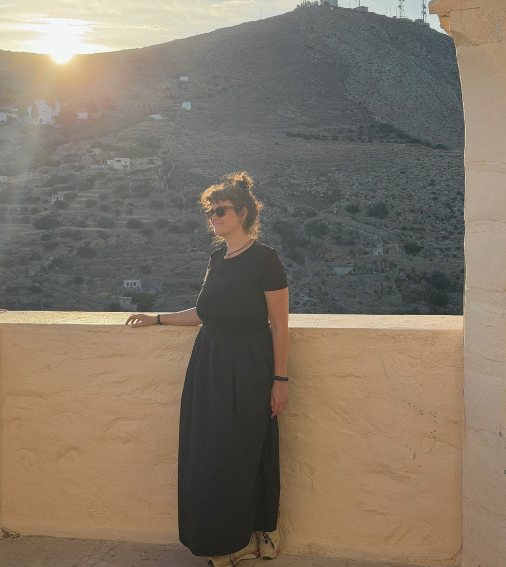 A woman with curly hair, wearing sunglasses, a black T-shirt, and a long black skirt, stands on a balcony during sunset with a mountain and scattered buildings in the background.